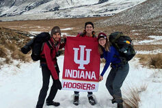 3 students hold an IU flag in front of snow-covered mountains.